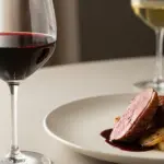 Sliced duck breast with sauce on a plate next to a wine glass on a table