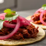 Tacos topped with shredded meat, pickled red onions, and cilantro leaves on a gray surface