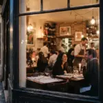 A couple dining at a table inside a warm, glass-walled bistro on a rainy, cobblestone street at dusk