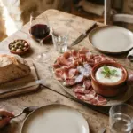 A rustic table holds a wine glass, bread, charcuterie, cheese, and olives set for a meal